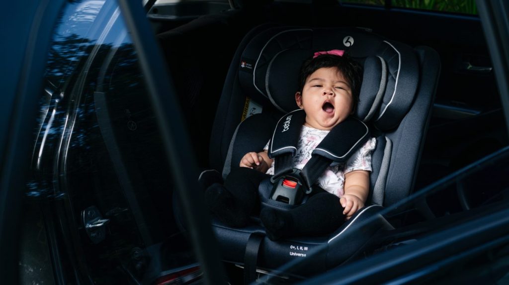 a baby girl is yawning while being strapped to a baby car seat.